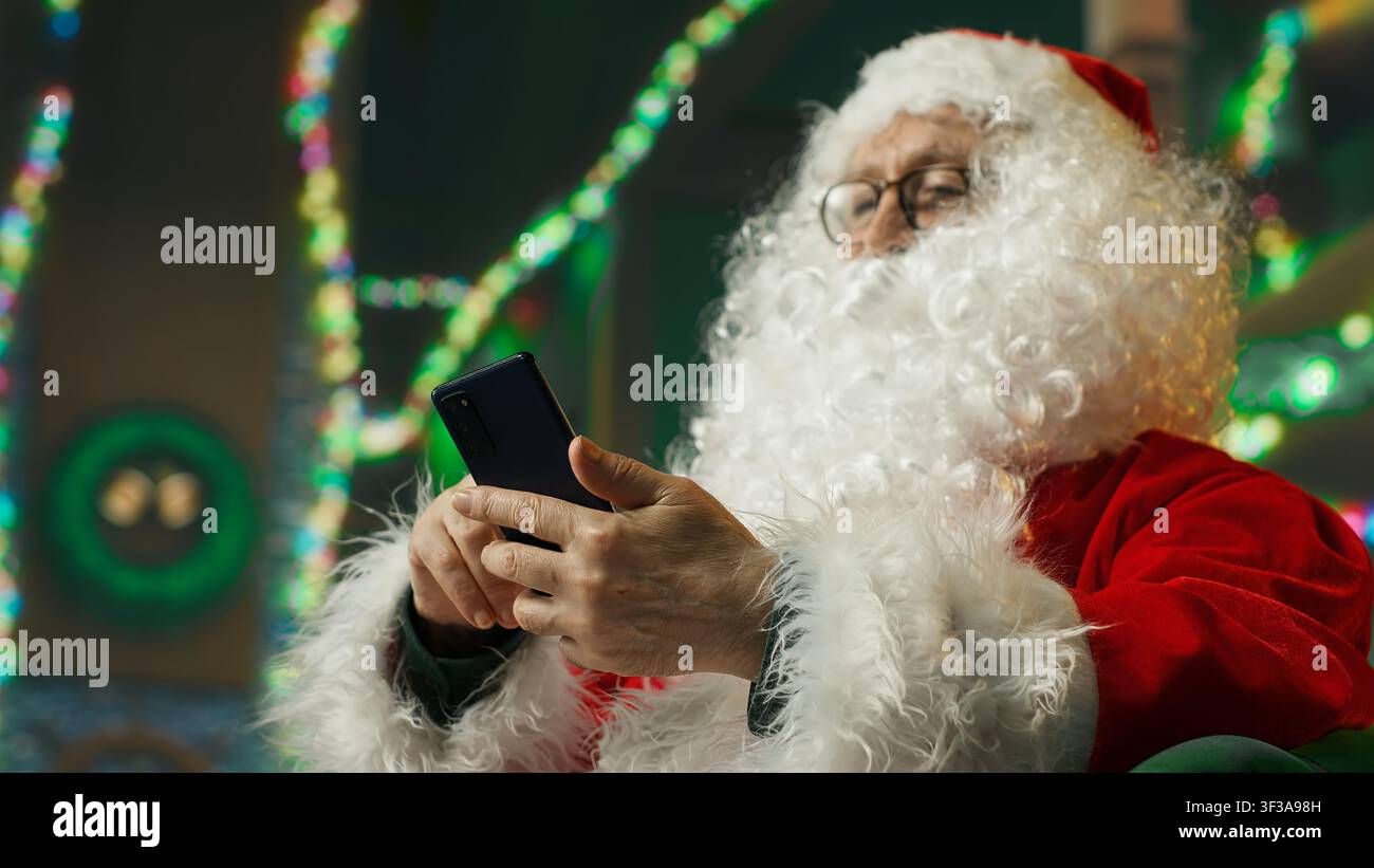 Glücklicher Mann, der als Weihnachtsmann gekleidet ist und lacht, während er lustige Videos auf dem Handy ansieht. Fröhliche Person, die Weihnachtskostüm trägt, amüsiert von Social Media Posts auf dem Telefon, Kamera Und Nahaufnahme Stockfoto