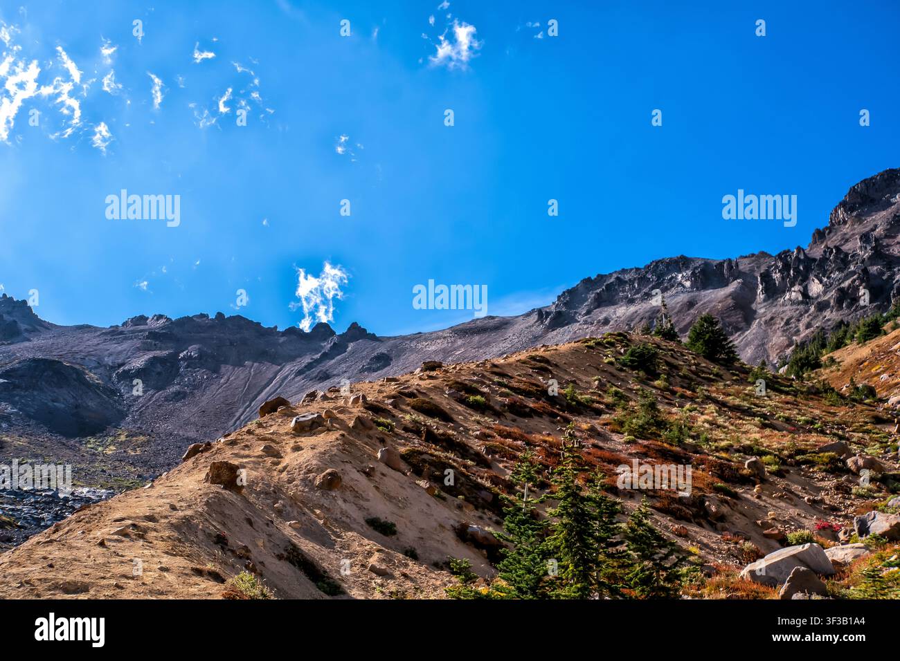 Schroffe Berge erheben sich unter einem lebendigen blauen Himmel. Spärliche Vegetation säumt das felsige Gelände. Wenige Wolken treiben träge in der Weite. Die Sonne wirft Stockfoto