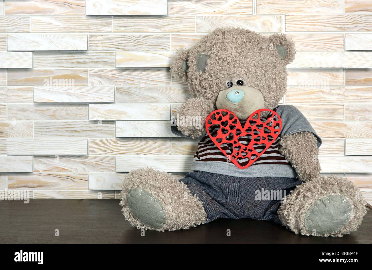 Ein Teddybär hält ein rotes Holzherz in seinen Pfoten. Ein Spielzeugbär sitzt auf einem Holzregal. Stockfoto