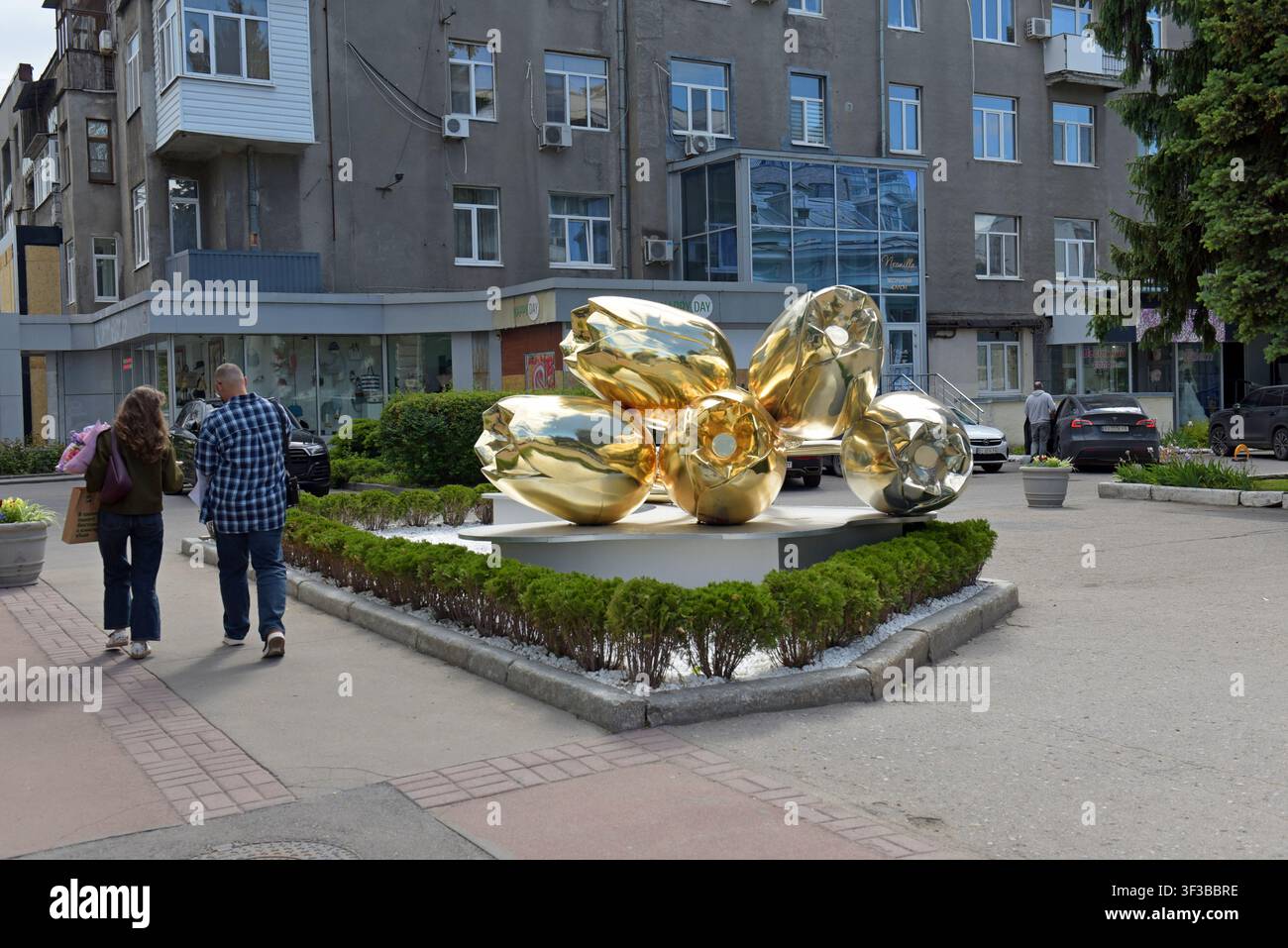 Menschen, die am Standesamt vorbeigehen, einem Ort für standesamtliche Hochzeiten mit einer Skulptur aus goldenen Tulpen draußen, in Charkiw, Ukraine, Mai 2026 Stockfoto