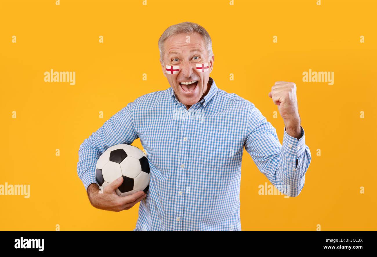 Glücklicher älterer kaukasischer Mann, der mit einem Fußball jubelt, mit England-Gesichtsbemalung, gelber Hintergrund Stockfoto