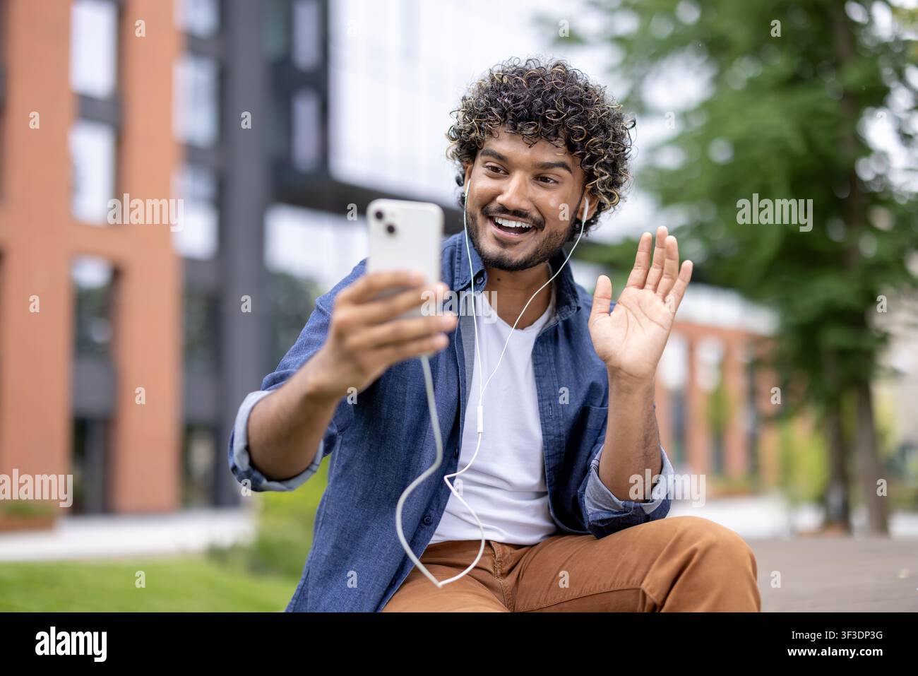 Lächelnder junger Mann mit lockigem Haar und Kopfhörern führt ein lebhaftes Videogespräch auf seinem Handy im Freien, winkt der Person auf dem Bildschirm zur Begrüßung zu und genießt die drahtlose Kommunikation Stockfoto