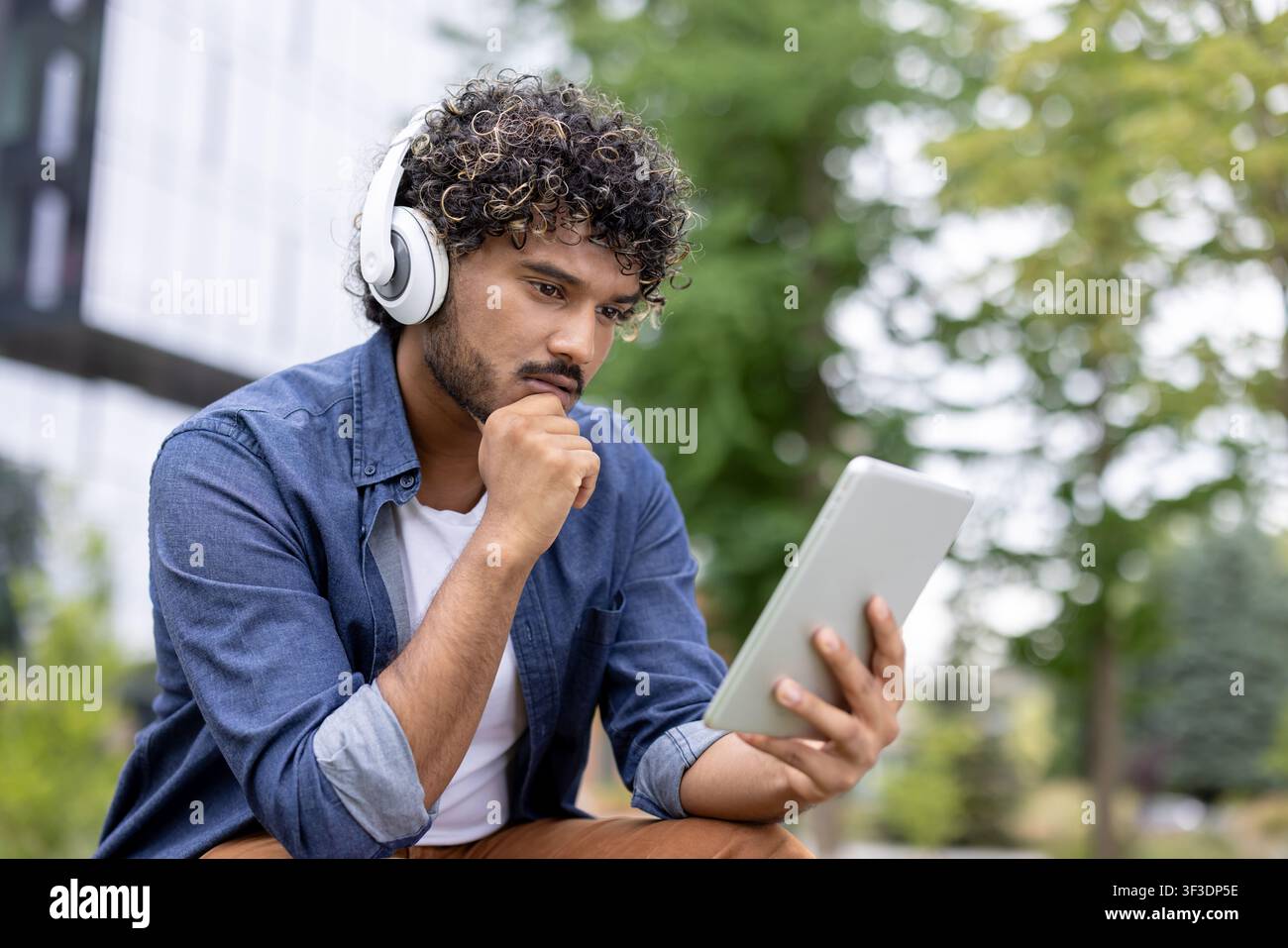 Junger nachdenklicher Mann mit Kopfhörern und lässiger Kleidung, konzentriert auf ein digitales Tablet im Freien, repräsentiert modernen Lebensstil, Technologie und Vernetzung Stockfoto