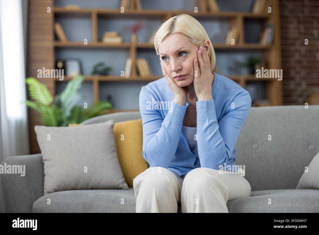 Reife Frau sitzt auf einem bequemen grauen Sofa in ihrem Wohnzimmer, legt die Hände an die Wangen, wirkt nachdenklich und reflektiert persönliche Herausforderungen oder emotionalen Stress Stockfoto
