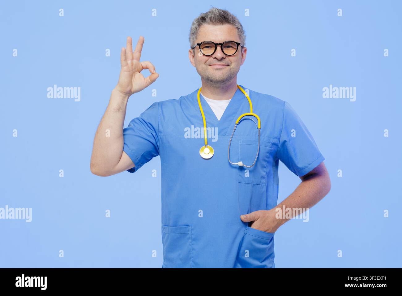 Ein selbstbewusster Arzt in OP-Kleidung macht ein Okay-Zeichen, das Zuversicht und Professionalität in einer modernen medizinischen Umgebung symbolisiert. Stockfoto