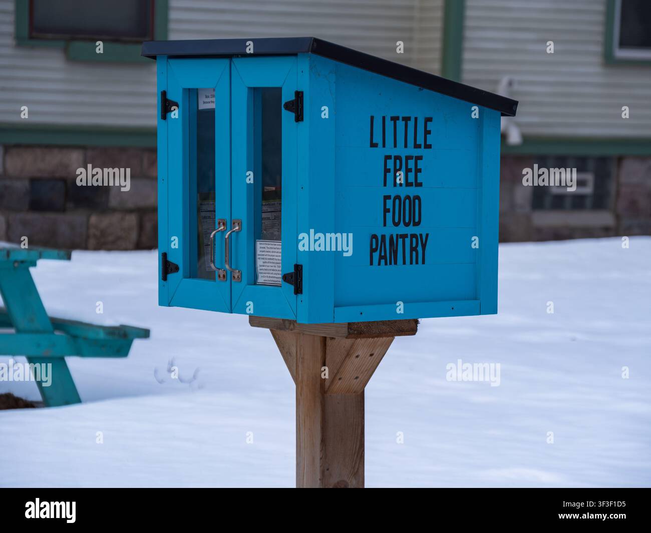 Helle blaue kleine Freigabeküche an einem Holzpfosten in einer verschneiten Nachbarschaft, die Gemeinschaftsunterstützung, Wohltätigkeit und Nahrungsmittelteilung im Winter zeigt. Stockfoto