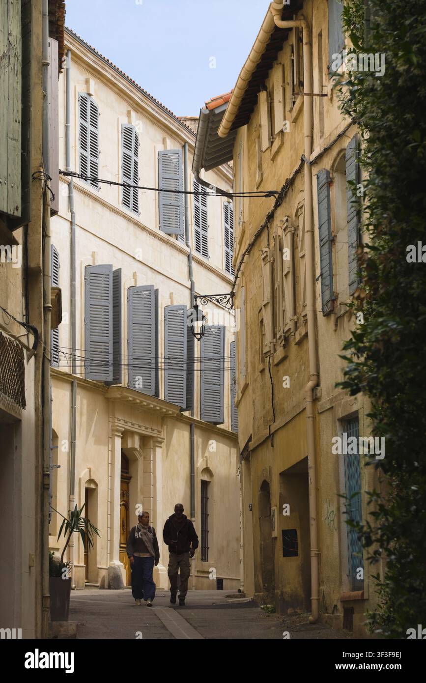 Zwei Fußgänger schlendern eine ruhige, schmale Straße entlang, die von hohen Steinbauten mit grauen Holzläden in Arles, Frankreich, gesäumt ist. Stockfoto