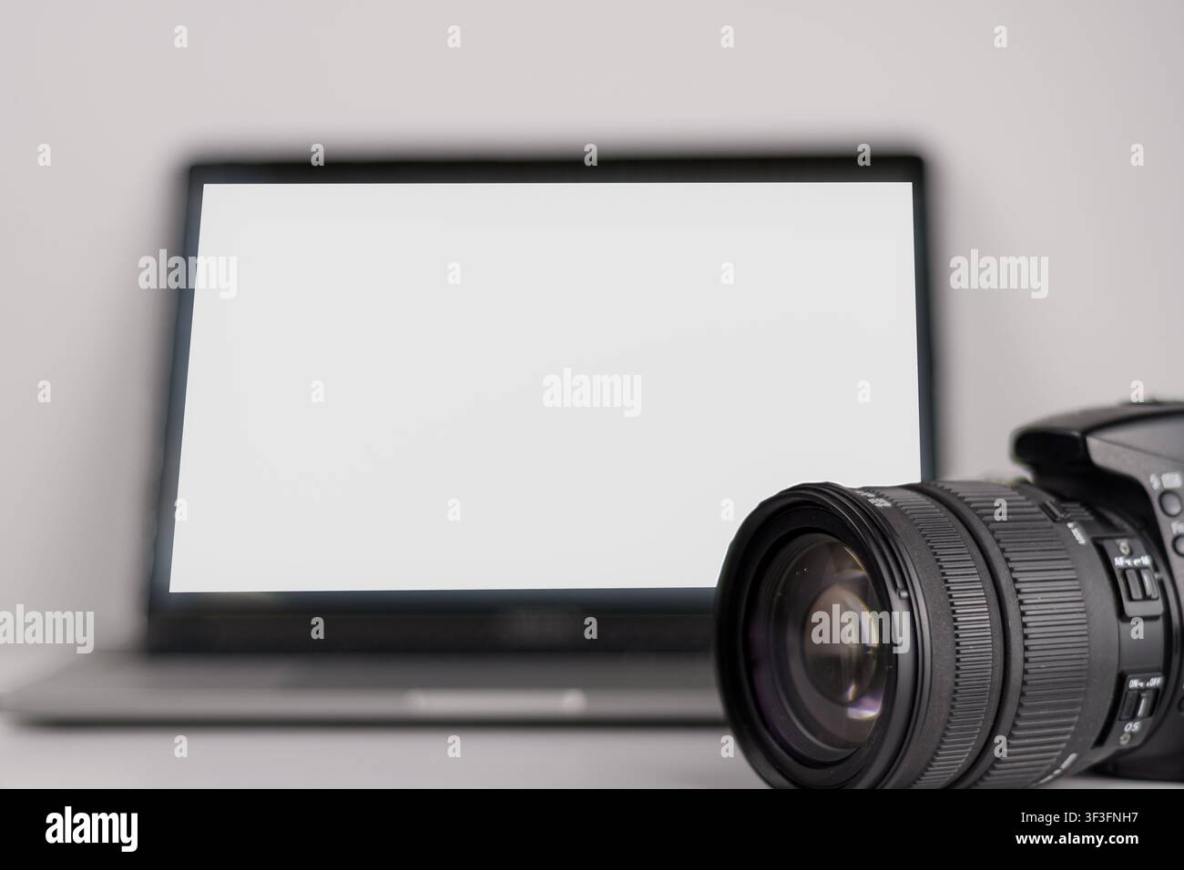 Nahaufnahme einer Digitalkamera und eines Objektivs neben einem Laptop mit leerem weißem Bildschirm, ein minimalistisches Arbeitsplatz-Modell für Fotografie, Bildbearbeitung, Portfolio und c Stockfoto