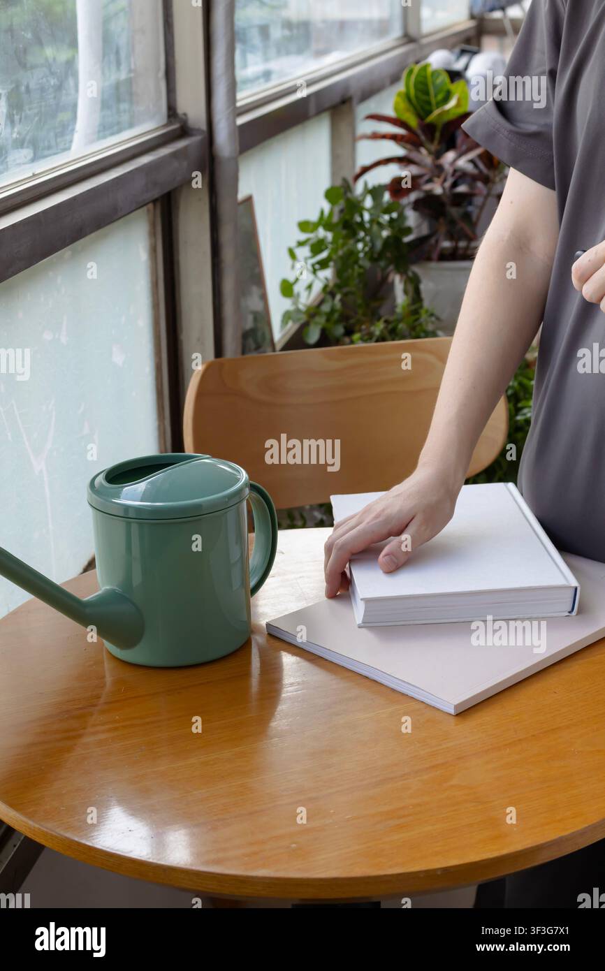 Hand, die ein leeres Hardcover-Buch auf einem Holztisch neben einem grünen Gießkanne in einem modernen Zuhause platziert Stockfoto