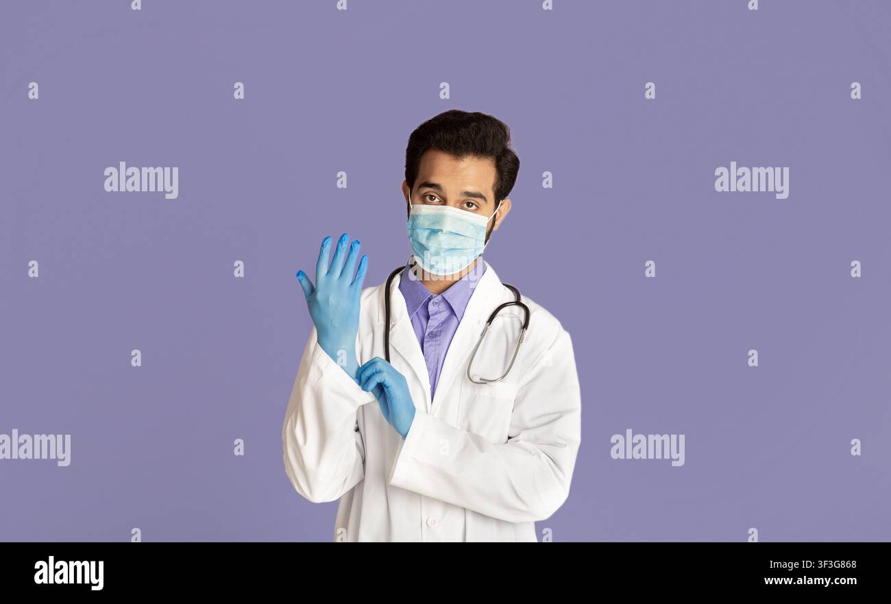 Schutz vor Viruserkrankungen. Indischer Arzt trägt medizinische Maske, Laborkittel und Gummihandschuhe auf violettem Hintergrund Stockfoto