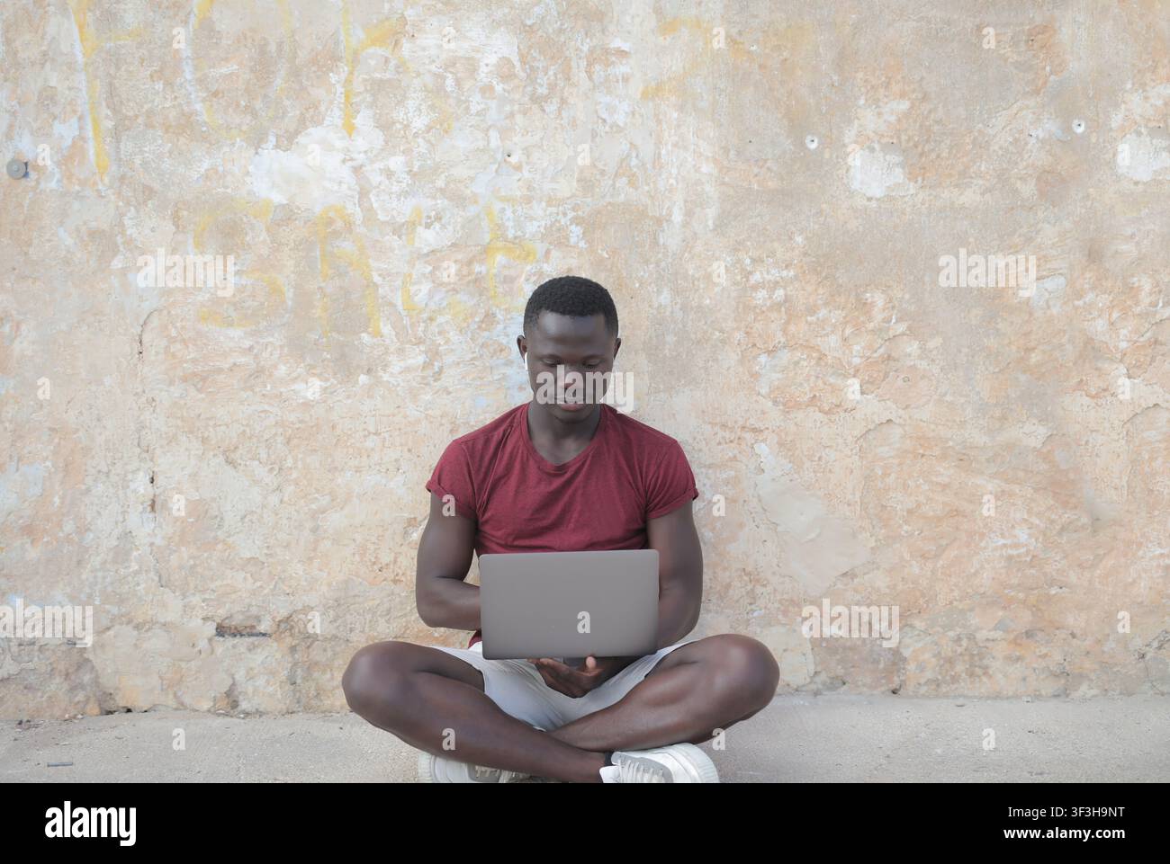 Junger Mann sitzt draußen mit Laptop und Ohrstöpseln Stockfoto