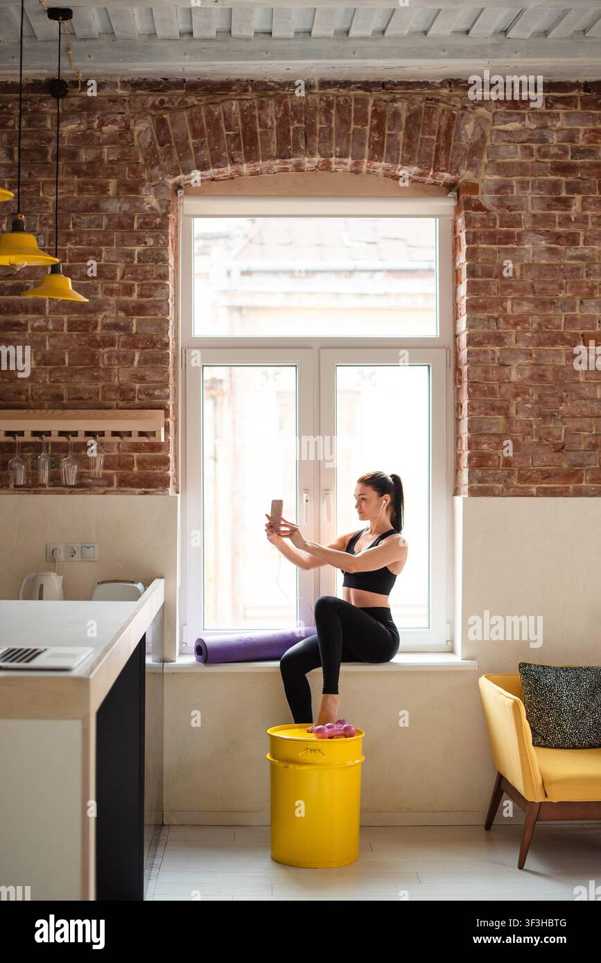 Sportliche Frau sitzt auf der Fensterbank und benutzt ein Smartphone Stockfoto