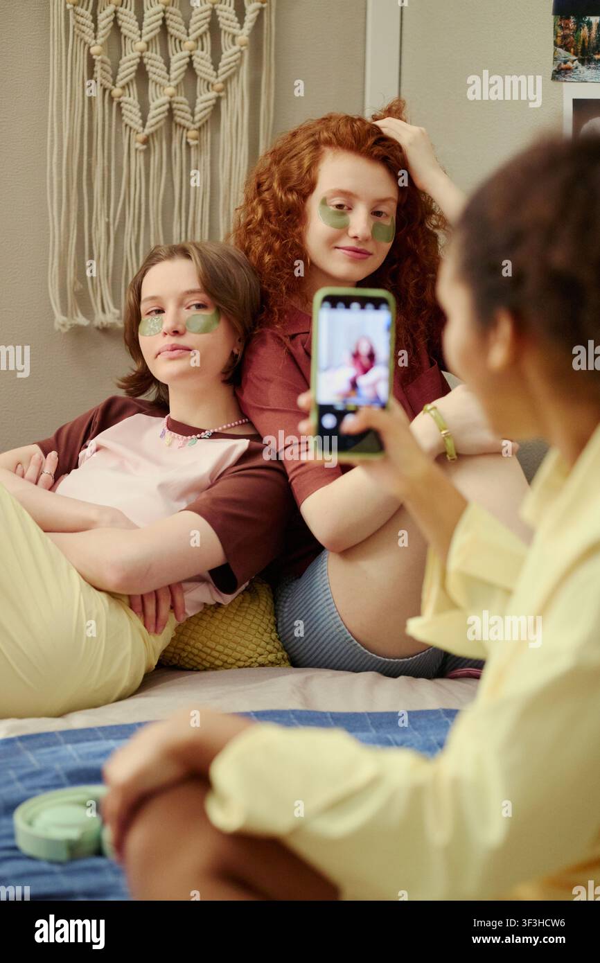 Vertikale Aufnahme von vielfältigen jungen Frauen, die mit Augenpflastern posieren, während eine Freundin den Hautpflegemoment mit einem Smartphone fotografiert und die Selbstpflege-Routine zeigt. Für Beauty-Marketing, Wellness-Inhalte, Social Media Stockfoto