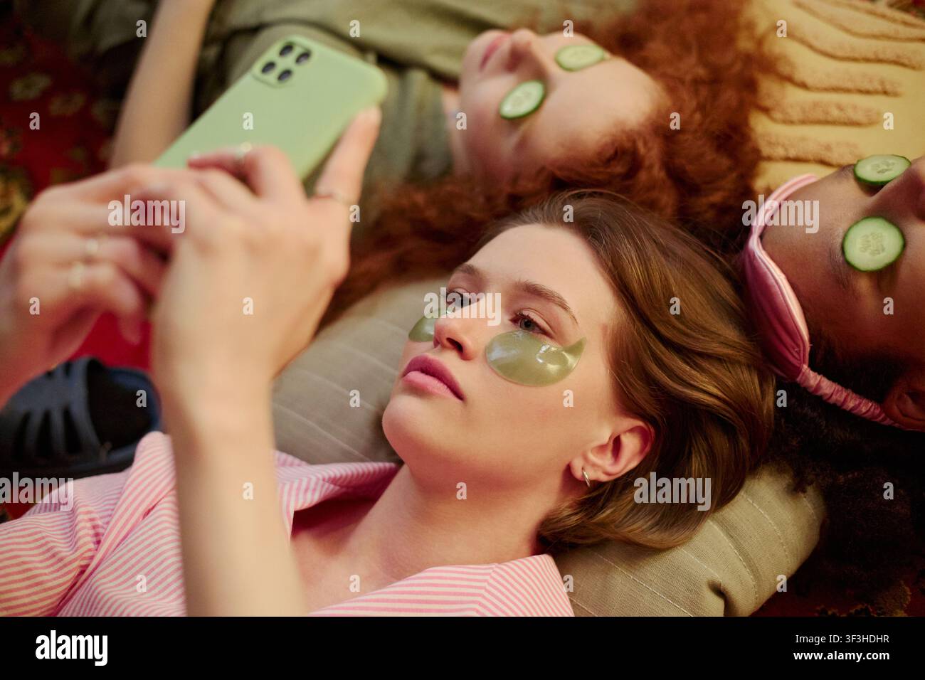 Junge Frauen entspannen mit Augenmasken und Gurkenscheiben, nutzen das Smartphone während einer Spa-Session zu Hause, zeigen Selbstfürsorge und Freundschaft. Ideal für Wellness, Schönheit, Lifestyle-Marketing Stockfoto