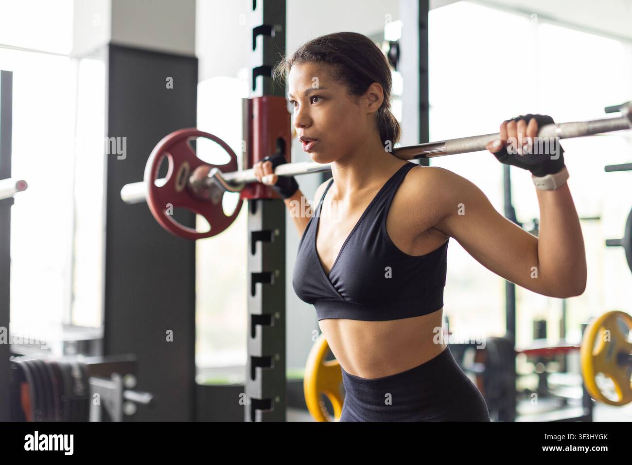 Junge schwarze Frau hält eine Hantelstange, während sie sich im Fitnessstudio zum Kniebeugen bereitmacht Stockfoto