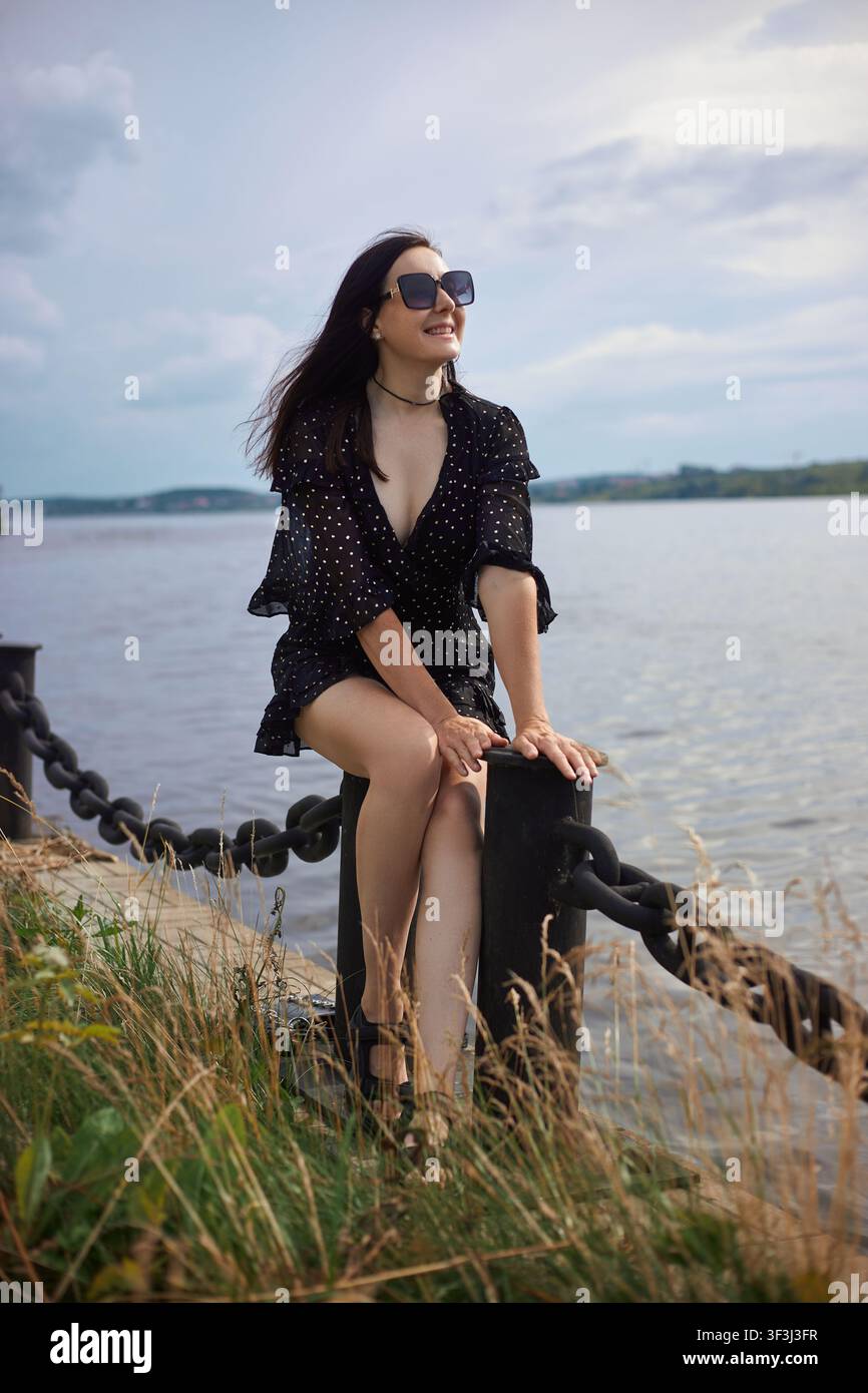 Frau mit langen Haaren sitzt an schwarzer Kette am Fluss. Sie trägt eine Sonnenbrille und ein schwarzes Kleid mit weißen Punkten. Der Himmel ist bewölkt und das Wasser ist ruhig im Hintergrund Stockfoto