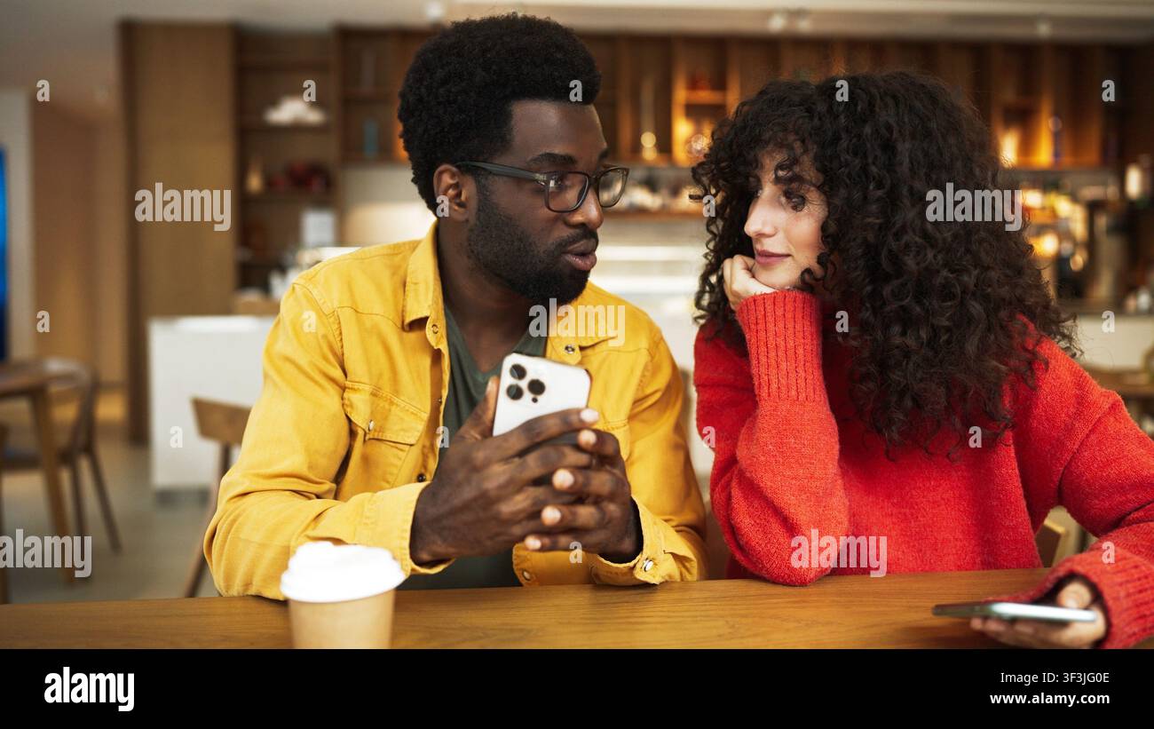 Mann in gelbem Hemd zeigt Smartphone einer Frau in rotem Pullover am Cafétisch. Afroamerikanischer Mann mit Brille hält Telefon, während lockighaarige Stockfoto
