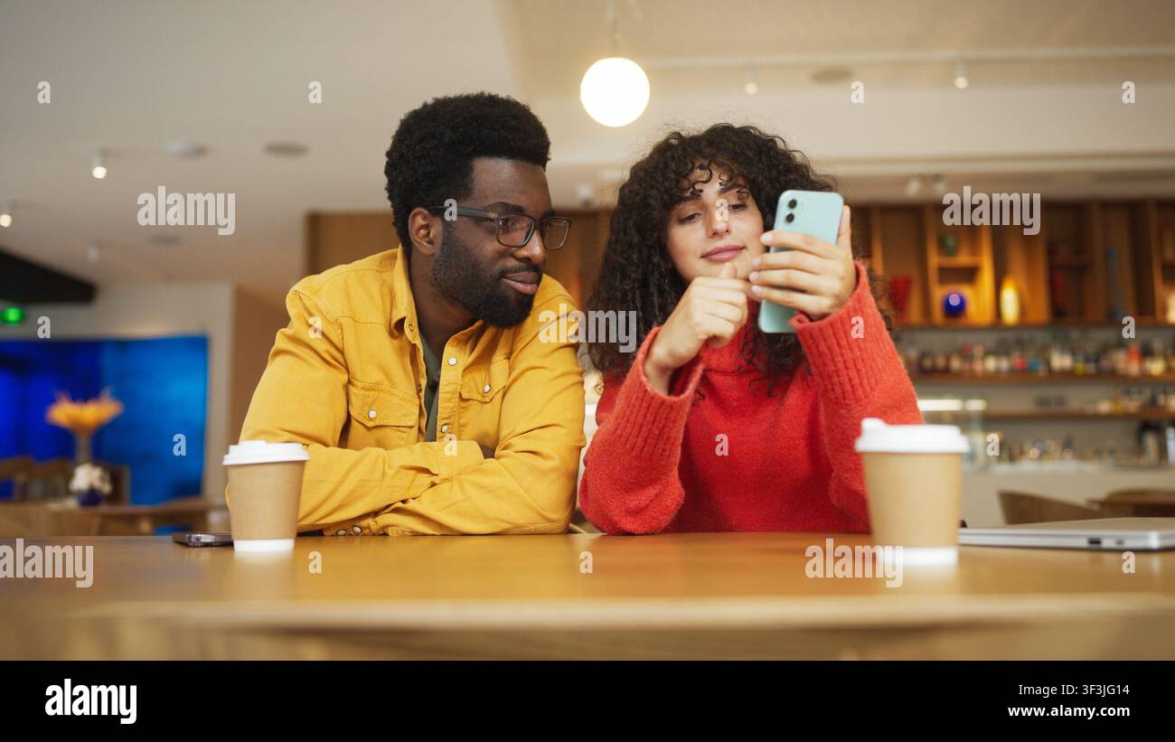 Mann mit gemischter Herkunft in gelbem Hemd und Frau in rotem Pullover sitzen an einem Café-Tisch mit Kaffeetassen und schauen gemeinsam auf ein Smartphone. Mann mit Brille Stockfoto