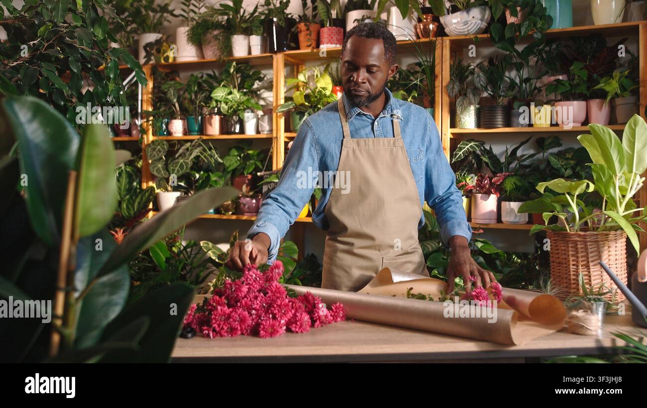 Fröhlicher professioneller Florist, der einen schönen Blumenstrauß in einem gemütlichen Blumenladen macht und dekoriert, schaut in die Kamera und lächelt. Schöne Afrikanerin Stockfoto