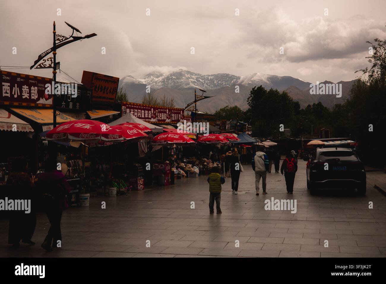 Blick auf eine belebte Straße in Lhasa mit roten Regenschirmen, Fußgängern, die gehen, und schneebedeckten Bergen im Hintergrund in Lhasa, Tibet, China. Stockfoto
