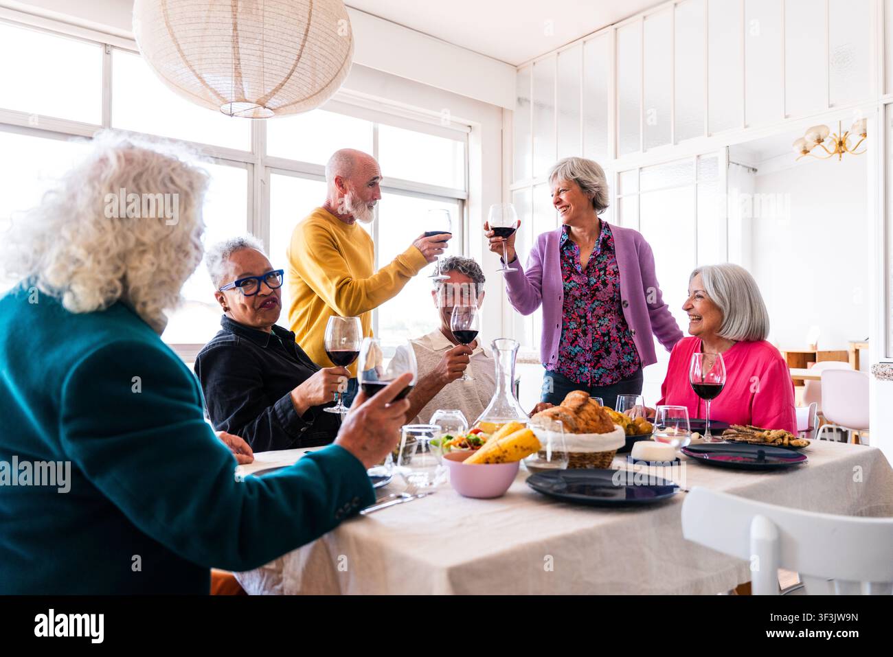 Gruppe lächelnder Seniorenfreunde, die eine fröhliche Dinnerparty zu Hause genießen, mit Rotwein anstoßen und Momente der Freundschaft und des Glücks teilen Stockfoto