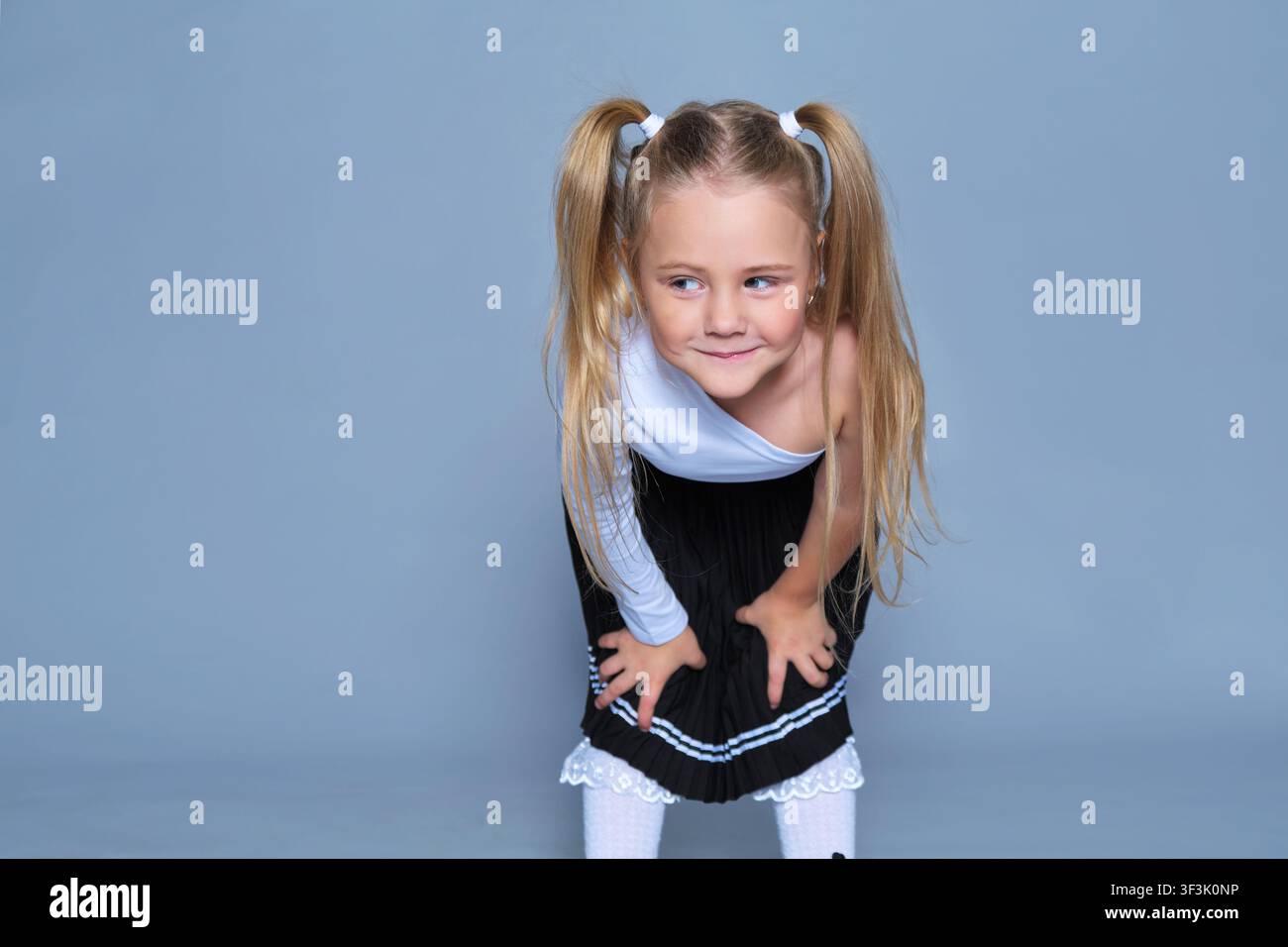 Verspieltes 5-jähriges Mädchen posiert in einer lustigen Haltung während eines Studio-Shoots, lehnt sich nach vorne mit einem schelmischen Lächeln. Gekleidet in einem modischen One-Shoulder-Top Stockfoto