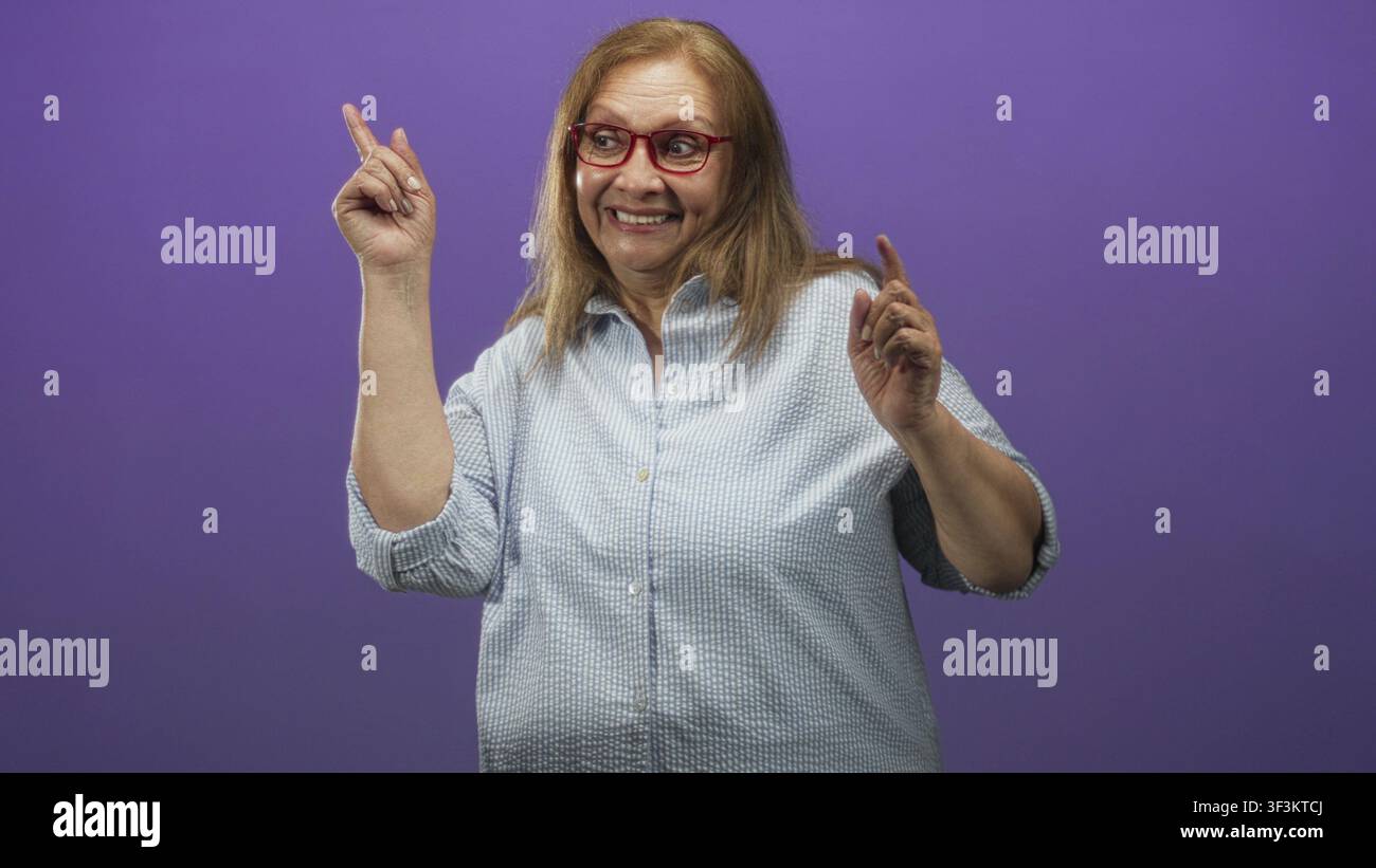 Seniorin zeigt mit beiden Zeigefingern nach oben und lächelt breit, trägt eine rote Brille und ein helles kariertes Hemd im Studio; Freudefeier. Stockfoto