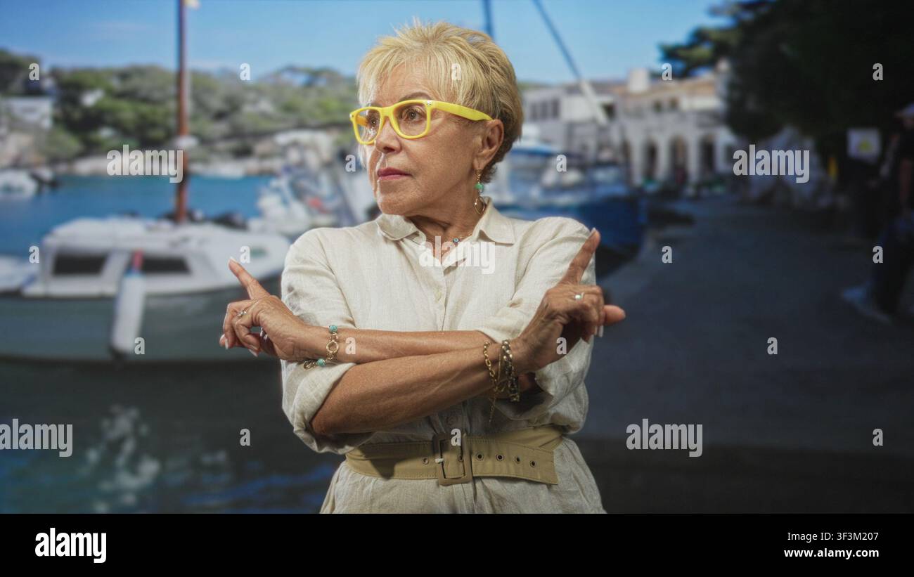 Ältere hispanische Frau mit gelber Brille und beigefarbenem Leinenkleid zeigt Zeigefinger mit gekreuzten Armen in einem Atelier mit Yachthafen-Hintergrund; feste diskrepanz Stockfoto