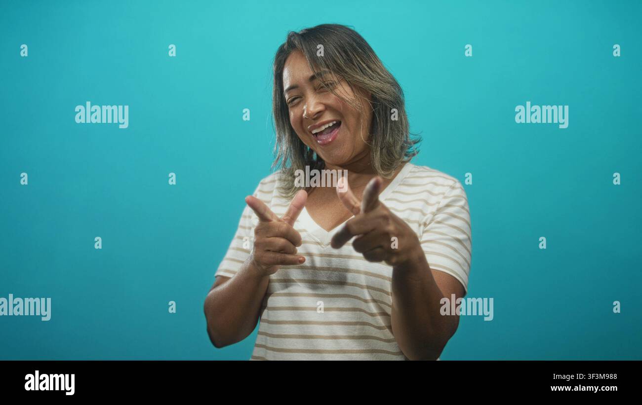 Frau zeigt beide Zeigefinger in Fingerpistolen, während sie im Studio mit türkisfarbener Kulisse lacht und lächelt; fröhliche Verspieltheit. Stockfoto