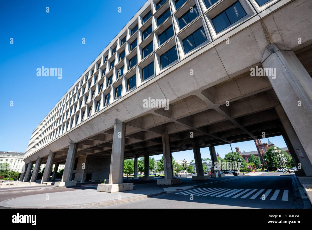 Forrestal-Gebäude auf Stelzen in Washington DC | WASHINGTON D.C., Vereinigte Staaten — Das James V. Forrestal-Gebäude spannt sich über die 10th Street SW auf massiven Betonpfeilern, seine brutalistische Masse rahmt einen entfernten Blick auf das Smithsonian Castle. Planer wollten einst diese Achse verbinden, um die modernistische Southwest-Neuentwicklung mit dem Smithsonian zu verknüpfen, aber das Navy Department baute dieses einzelne Gebäude über die Promenade statt zwei verbundener Türme, sodass heute nur noch die Spitze des Castle über der Dachlinie sichtbar ist. Stockfoto