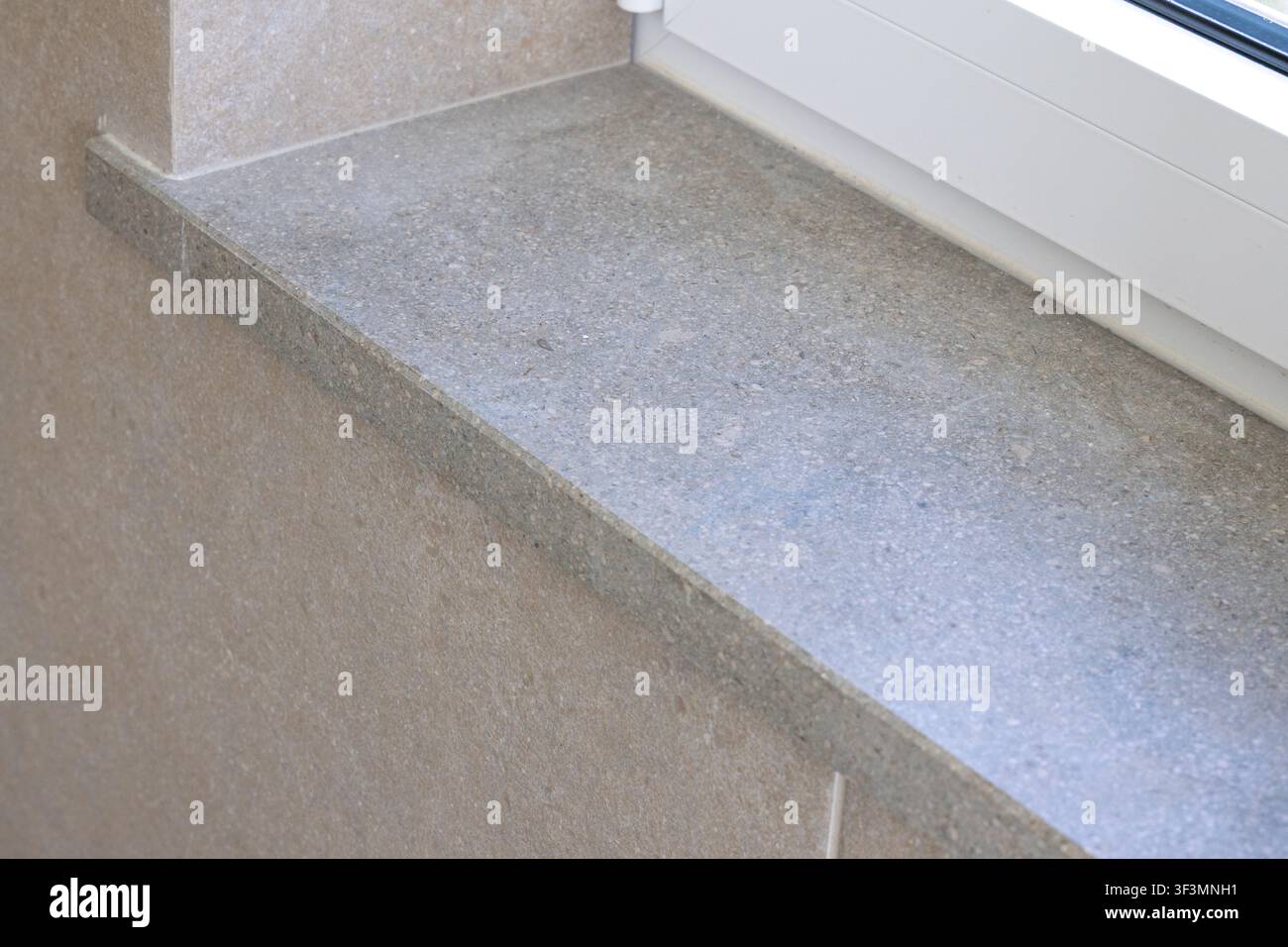 Ein detaillierter Schnappschuss zeigt die strukturierte Oberfläche einer grauen Granitfensterbank neben einer warmen beigen Fliesenwand und betont architektonische Details Stockfoto