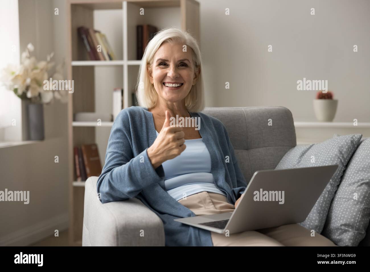 Seniorin hält Laptop, schaut in die Kamera und zeigt Daumen hoch Stockfoto