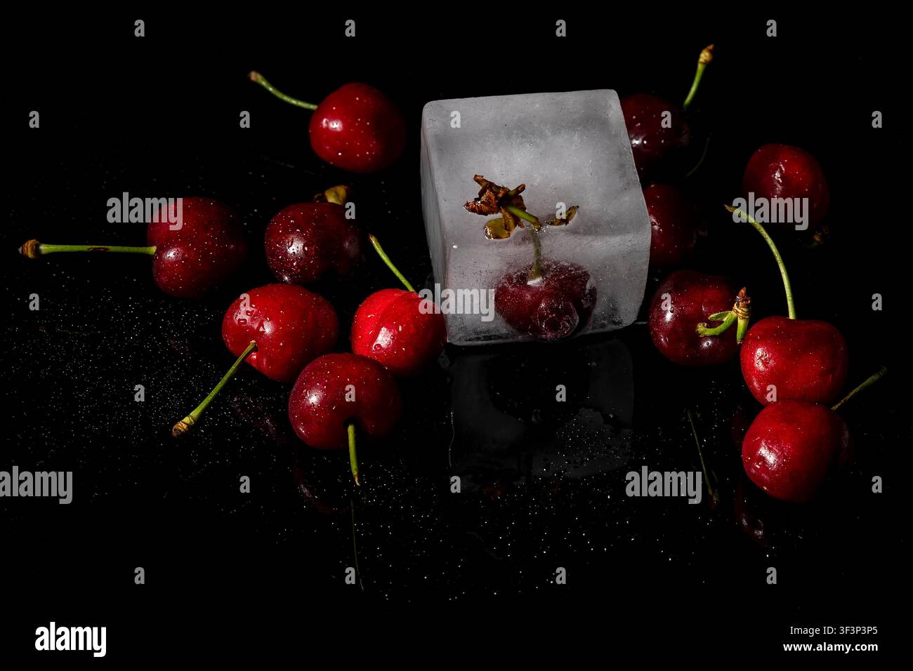 Eine Gruppe Kirschen ist mit Wassertropfen benetzt und eine ist in einem Eiswürfel gefangen, auf einem glänzenden schwarzen reflektierenden Hintergrund Stockfoto