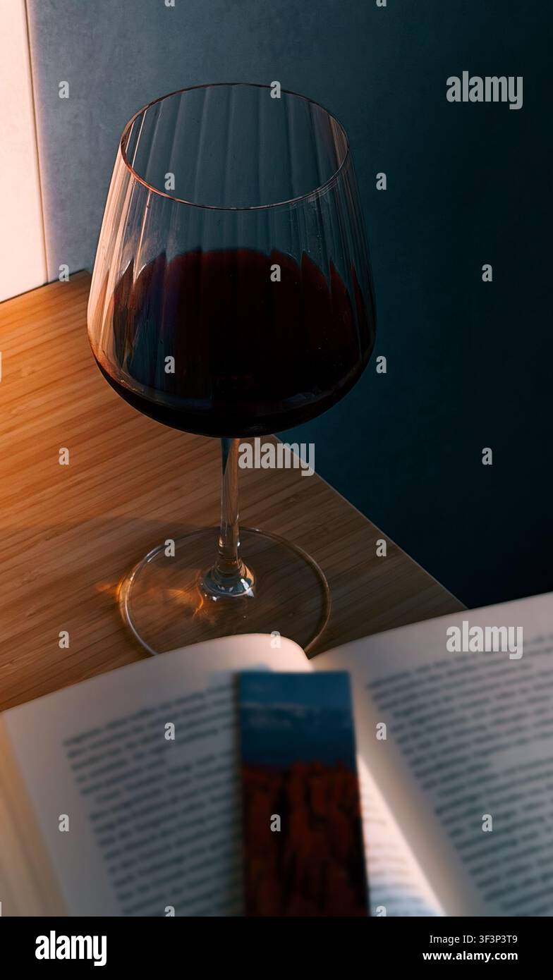 Glas Rotwein auf einem Holztisch neben einem offenen Buch mit Lesezeichen, weiches Licht beleuchtet die Szene von der Seite Stockfoto