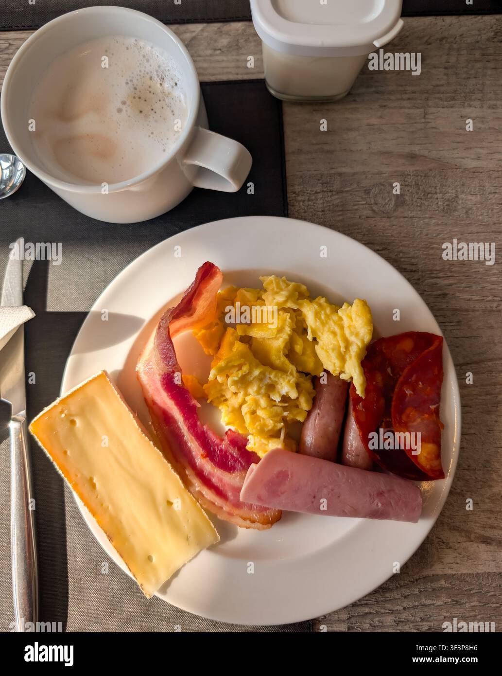Oberansicht eines herzhaften Hotelfrühstückstellers mit Rührei, Speck, Würstchen, Schinken, Käse und einer Tasse Cappuccino. Stockfoto