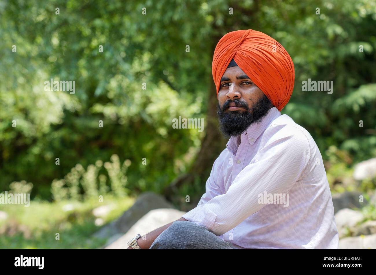 Nahaufnahme-Porträt eines jungen Sikh-Mannes mit einem orangefarbenen Turban, der direkt in die Kamera schaut, während er vor einem verschwommenen Hintergrund aus üppigem Grün sitzt Stockfoto
