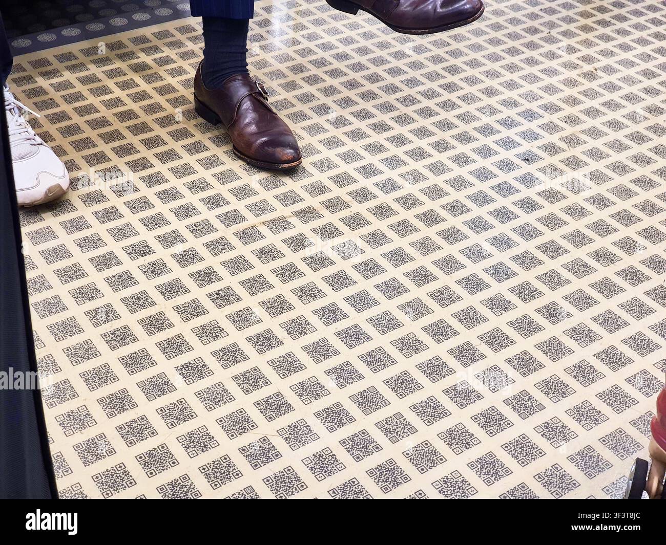 Eine Nahaufnahme eines Bodens, der vollständig mit einem sich wiederholenden Rastermuster aus QR-Codes bedeckt ist, die auf beigen Fliesen gedruckt sind. Das geometrische Schwarz-Weiß Stockfoto