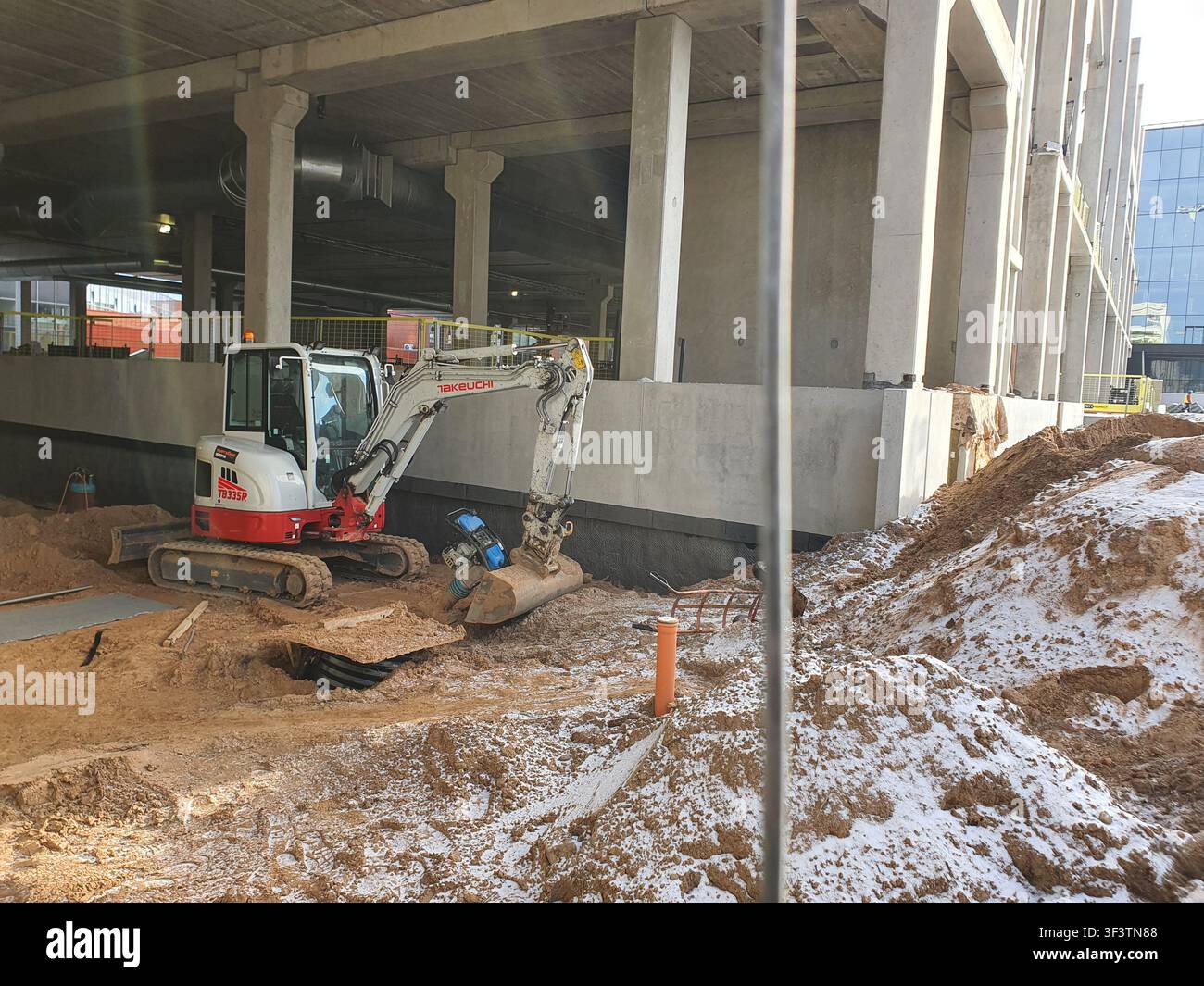 Ein kompakter roter und weißer Takeuchi TB235FR-Minibagger, der auf sandigem Boden auf einer Baustelle neben einer großen Betonbaustruktur positioniert ist Stockfoto