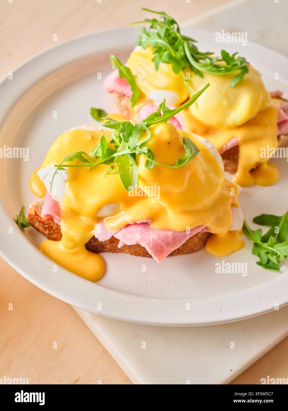 Ein köstlicher Teller Eier Benedikt, serviert mit cremiger Hollandaise-Sauce, Schinken und Rucola, perfekt für ein herzhaftes Frühstück. Auf einem weißen Teller aufgenommen für Stockfoto
