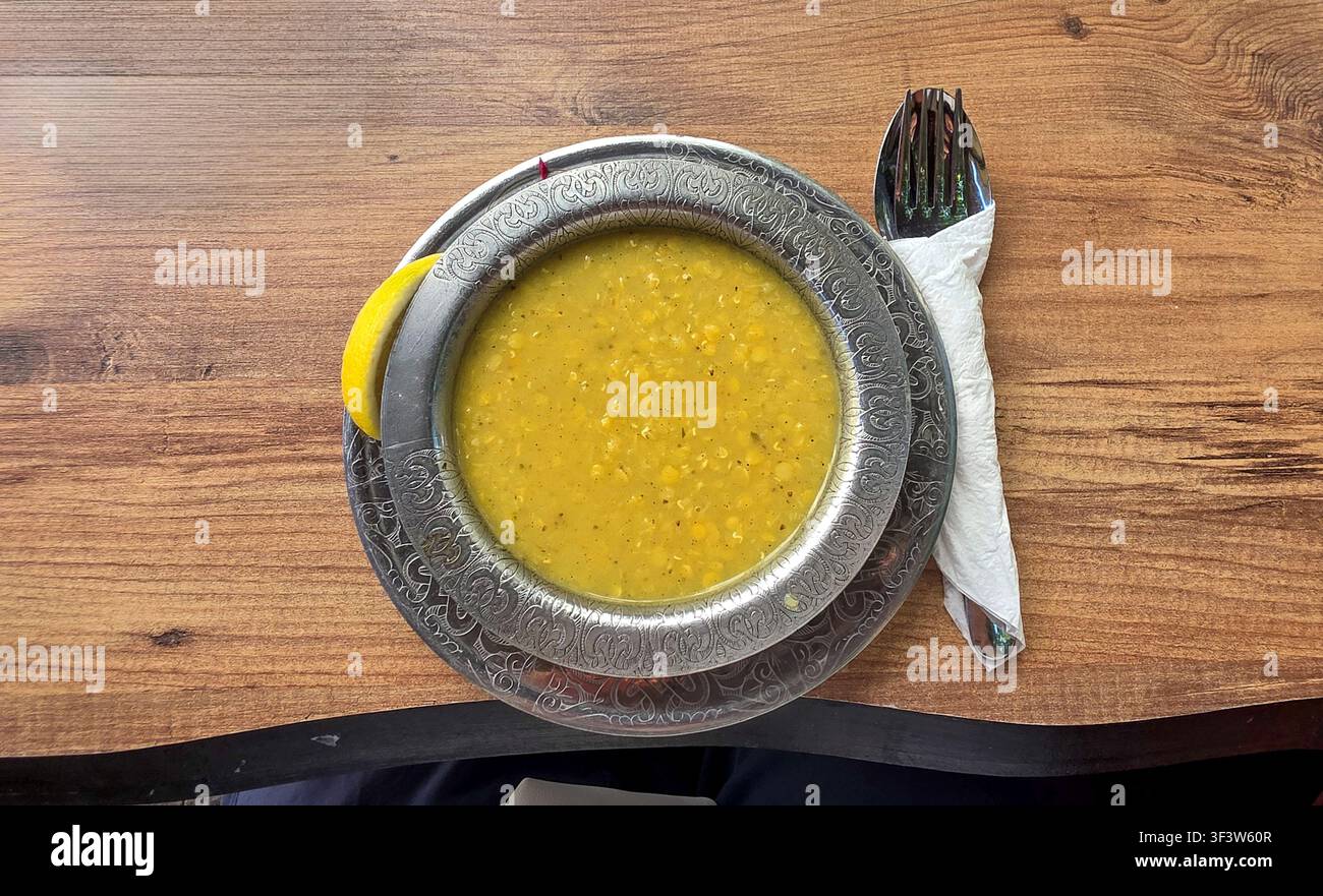 Eine warme, tröstliche gelbe Linsensuppe, serviert in einer dekorativen Metallschale mit aufwendigen Mustern, begleitet von einer Zitronenscheibe und Besteck auf Holz Stockfoto