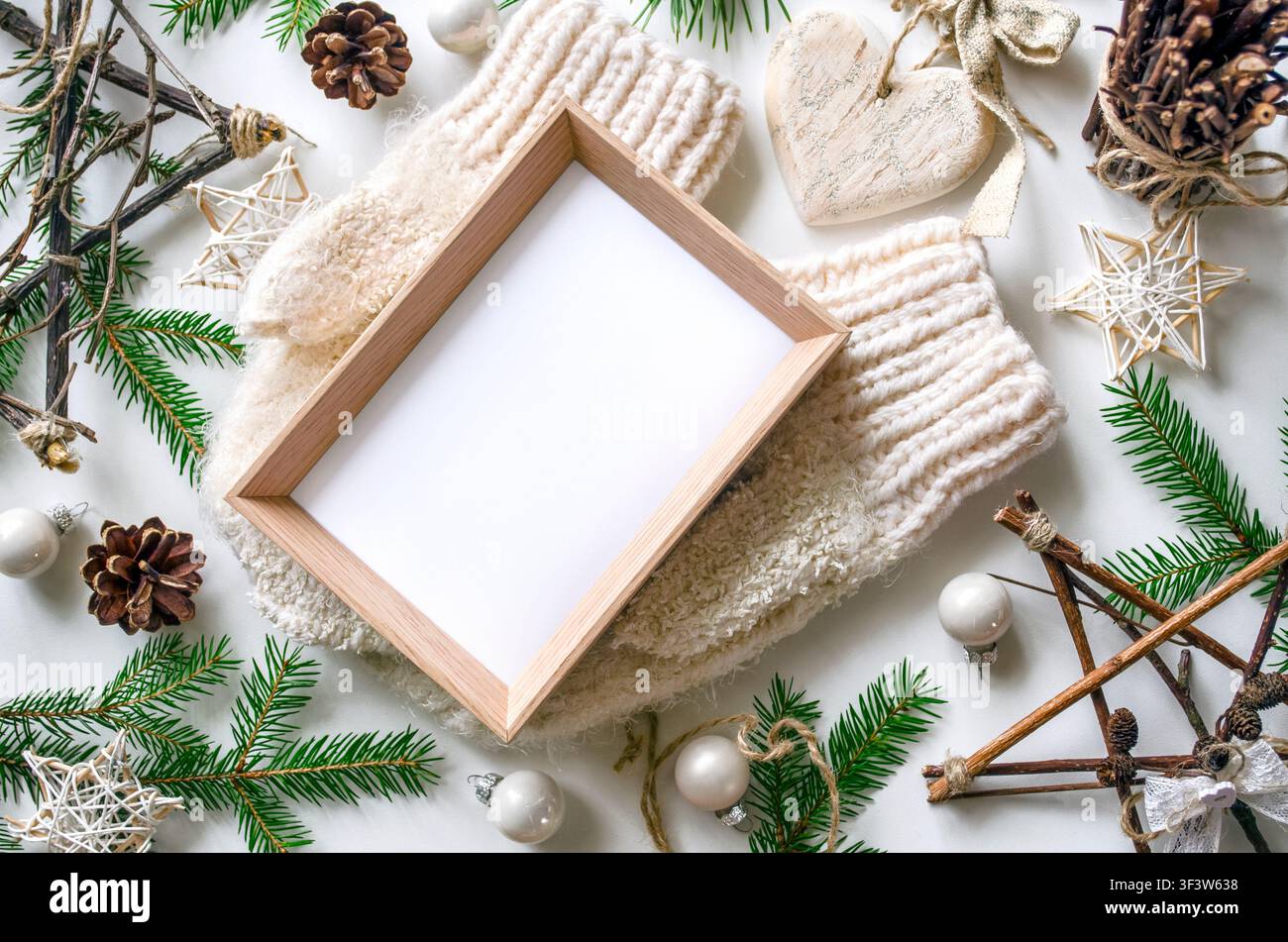 Gemütliches Weihnachts-Flatlay mit warmer gestrickter Faust, leerem Holzrahmen für Text, Tannenwedeln, Tannenzapfen, Sternornamente und Neujahrsdekoration auf weißem Hintergrund Stockfoto
