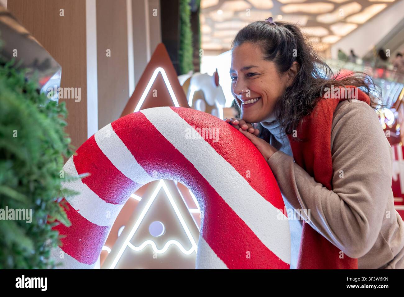 Lächelnde Frau lehnt sich an eine riesige rote und weiße Zuckerstangen-Dekoration auf einem festlichen Weihnachtsmarkt in China, mit leuchtenden Lichtern und fröhlicher Weihnachtsstimmung Stockfoto