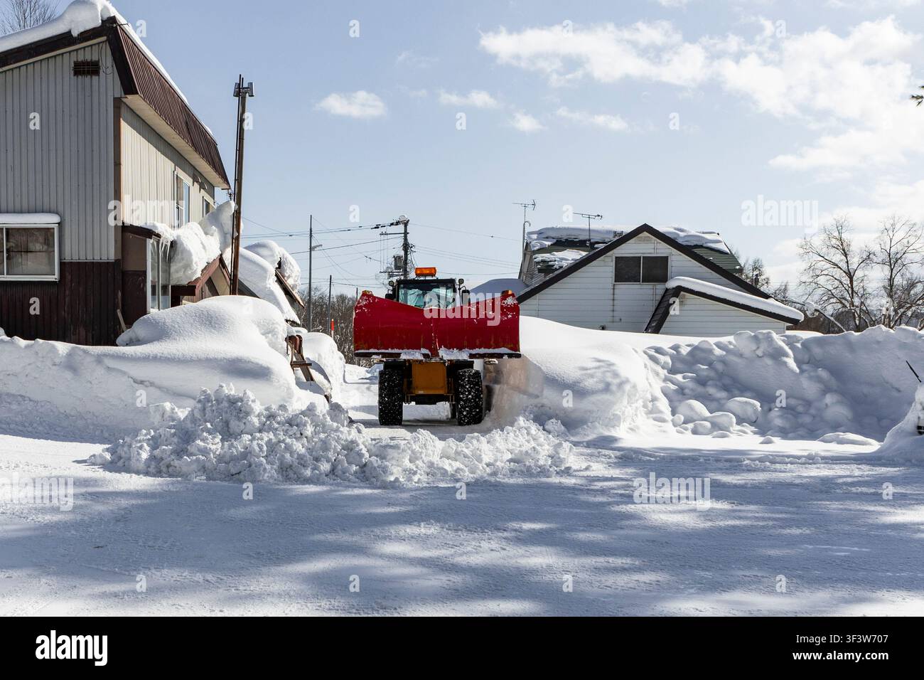 Ein kompakter Schneeräumtraktor schiebt frischen Schnee entlang einer Wohnstraße in Niseko, Hokkaido, Japan, umgeben von tiefen Schneeverwehungen, Häusern und hellem Stockfoto