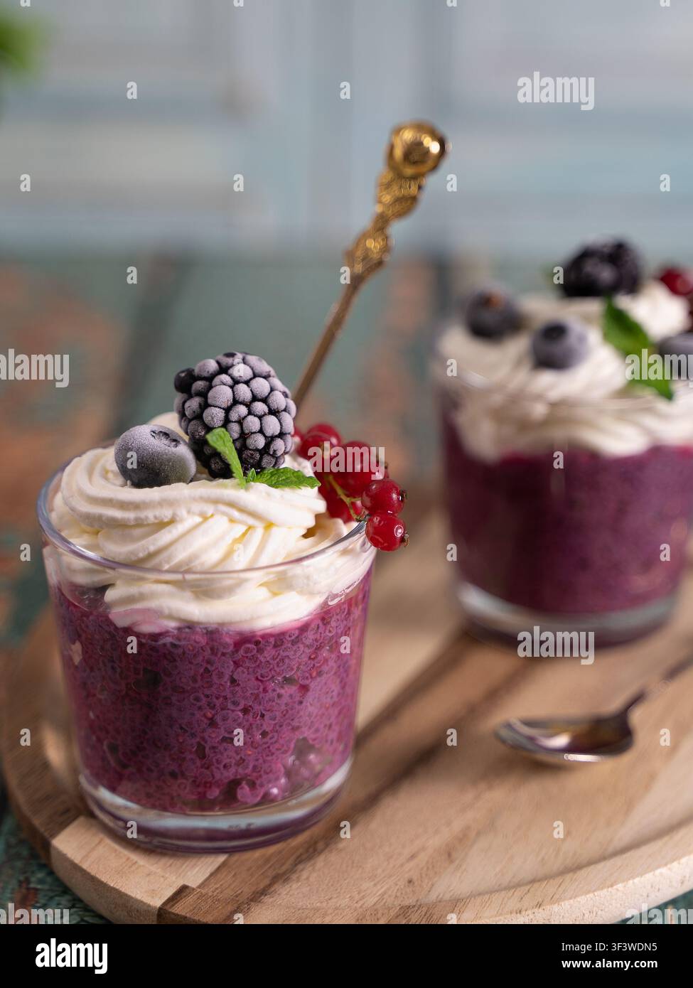 Vertikale Food-Fotografie eines geschichteten Beeren-Smoothie-Desserts, gekrönt mit Schlagsahne, Brombeere und roten Johannisbeeren auf einem rustikalen Holztisch Stockfoto