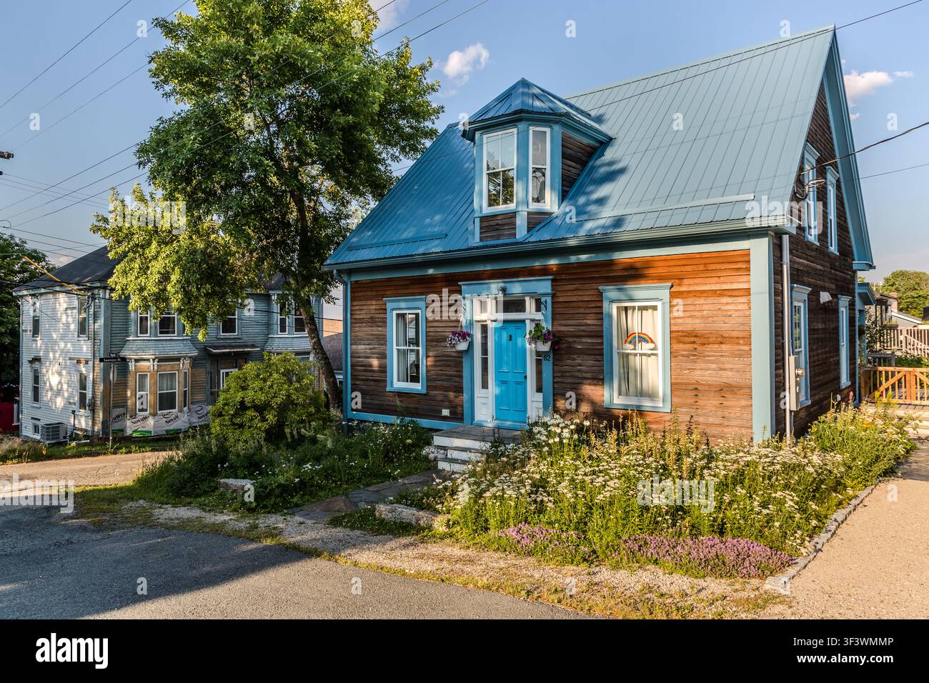 Ein braunes Holzhaus mit einer blauen Tür und blauem Dach steht in einer Wohnstraße in der Altstadt von Lunenburg, Nova Scotia, umgeben von einem Blumengarten. Die farbenfroh gestrichenen Holzhäuser kennzeichnen dieses historische Viertel, das seit 1995 UNESCO-Weltkulturerbe ist. Lunenburg, Nova Scotia, Kanada Stockfoto