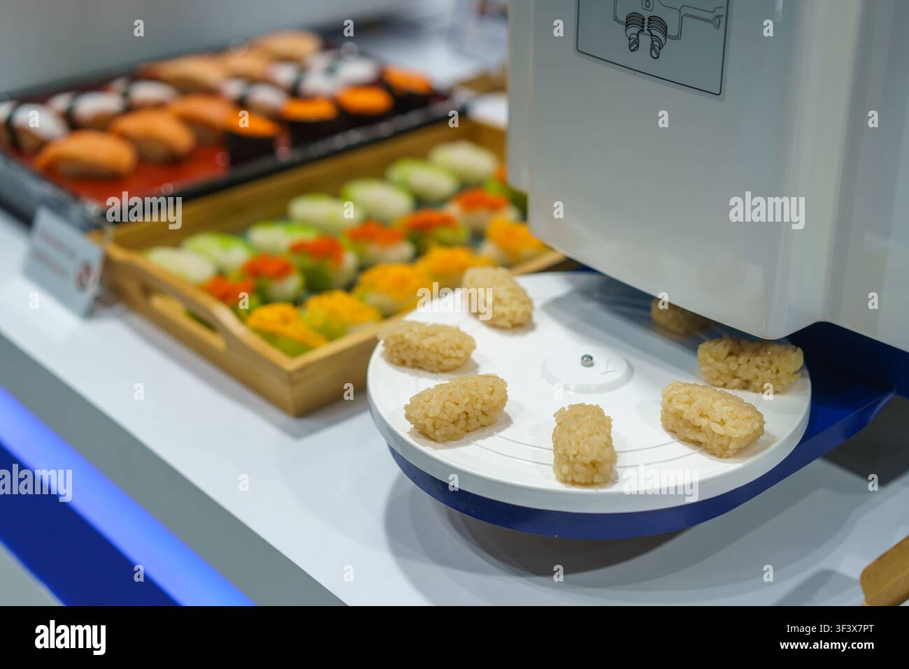 Nahaufnahme einer automatisierten kommerziellen Sushi-Maschine, die perfekt geformte Nigiri-Reiskugeln auf ein rotierendes Tablett abgibt, mit fertigem Sushi im Hintergrund Stockfoto