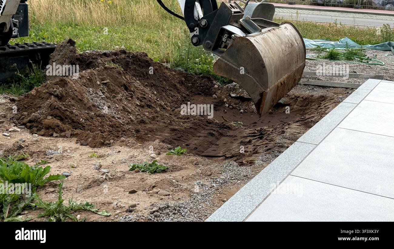 Baggerbagger gräbt in den Boden, mit einem Haufen Erde und einem gepflasterten Bereich im Hintergrund Stockfoto