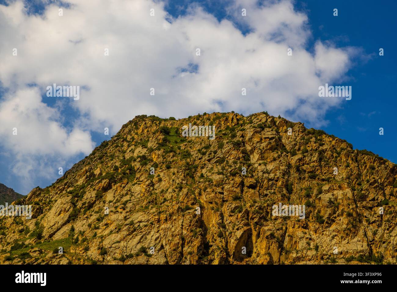 Felsiger Berghang zeigt verstreute grüne Vegetation bei hellem Tageslicht. Klarer Himmel mit vereinzelten Wolken unterstreicht die raue Naturlandschaft Stockfoto