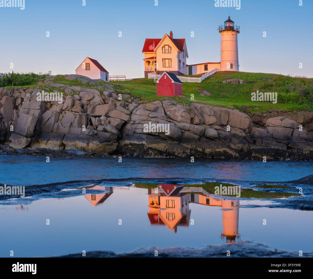Der Nubble-Leuchtturm steht an einer felsigen Küste mit einem Haus daneben. Die Strukturen spiegeln sich im ruhigen Wasser unten wider und schaffen eine ruhige Szene. Stockfoto