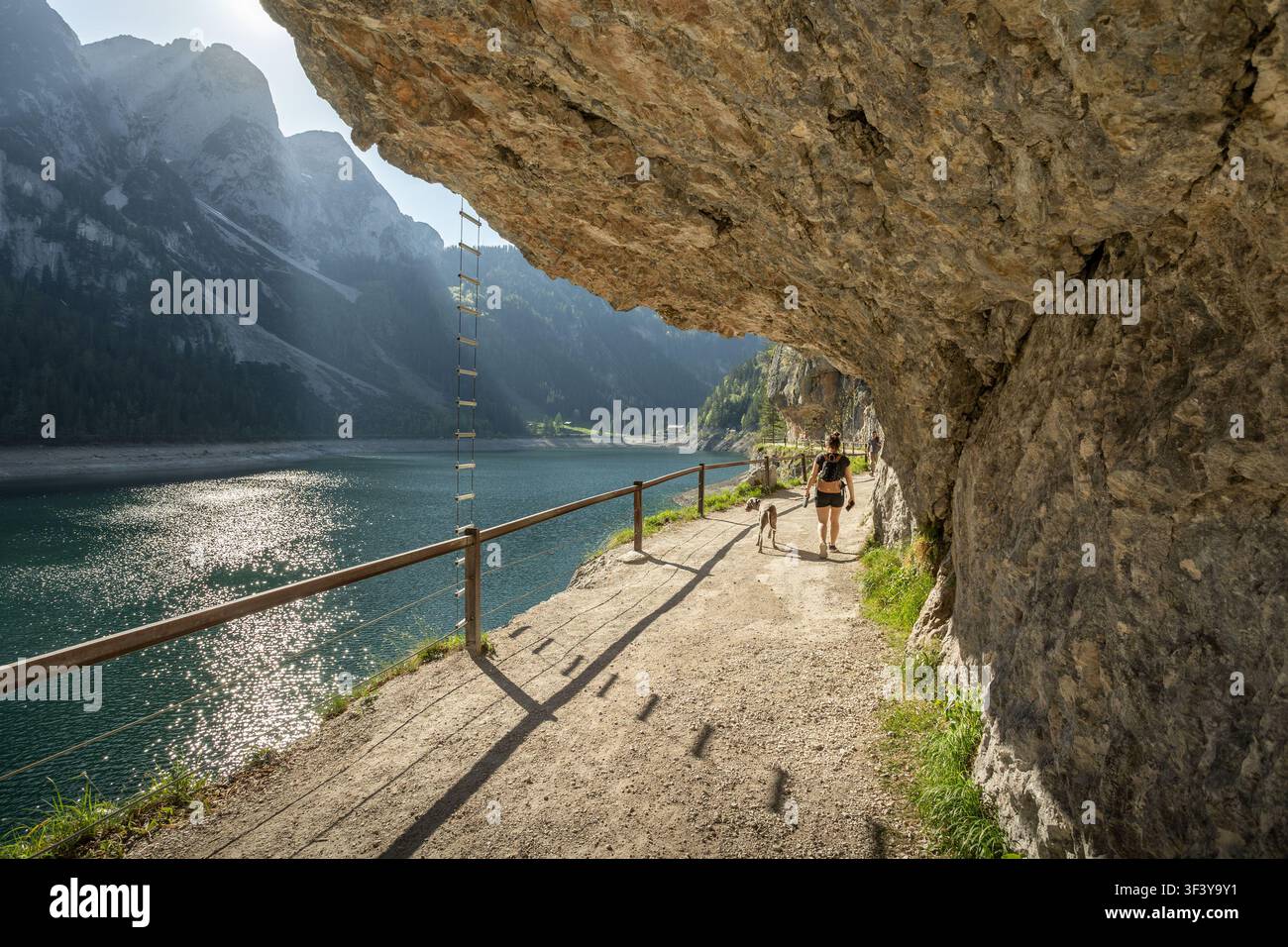 Wanderer und Hund gehen entlang des malerischen Pfades am Fore Gosau See unter einem Felsvorsprung Stockfoto